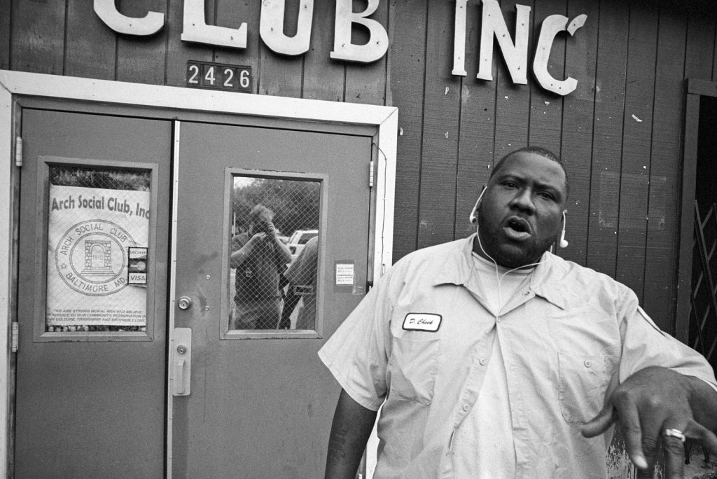 Black and white photo of a man in a button down shirt with headphones hanging from his ears stood outside Arch Social Club in Baltimore. He is pointing at the person taking the photograph, who can be seen with the camera reflected in a window on the buildings door.