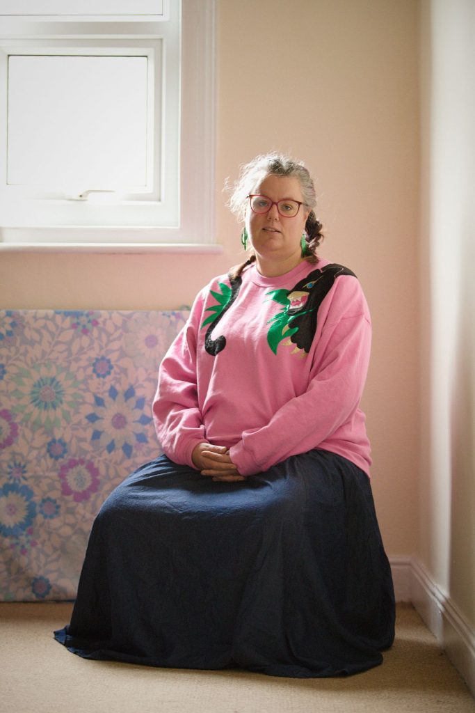 A woman sits on a stool in a light room with a window to the upper left. She is wearing a long navy skirt and a pink jumper with a panther around it.