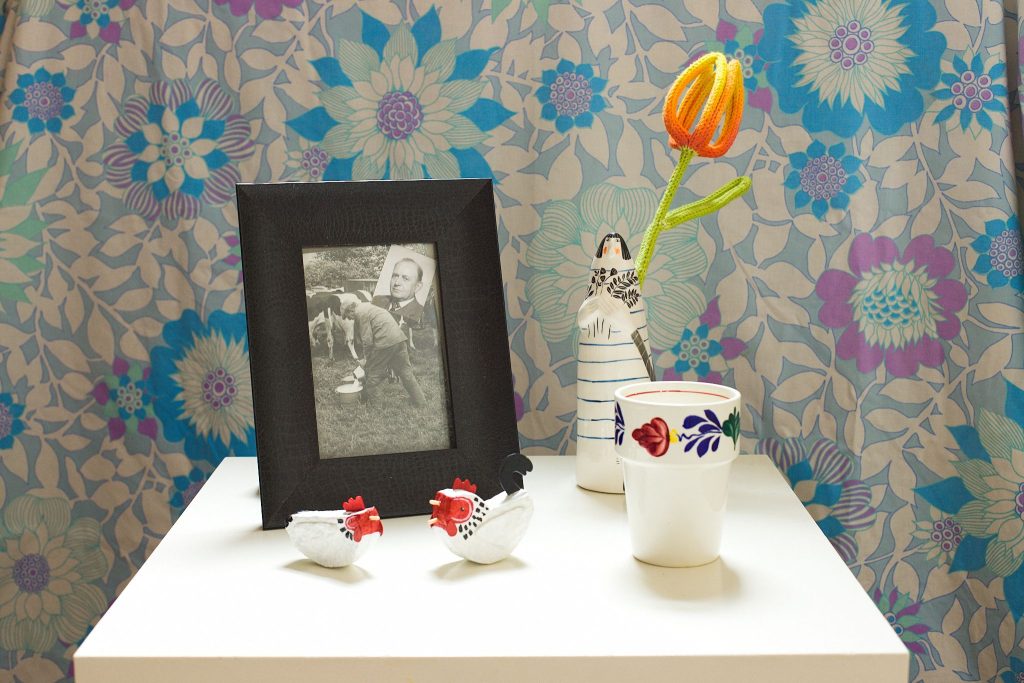 A white table with a series of objects sits in front of a blue patterned background. There are two small chickens, a black and white framed photograph of a man milking cows, a vase shaped like a woman with a woollen tulip in it and a white cup with a flower pattern.