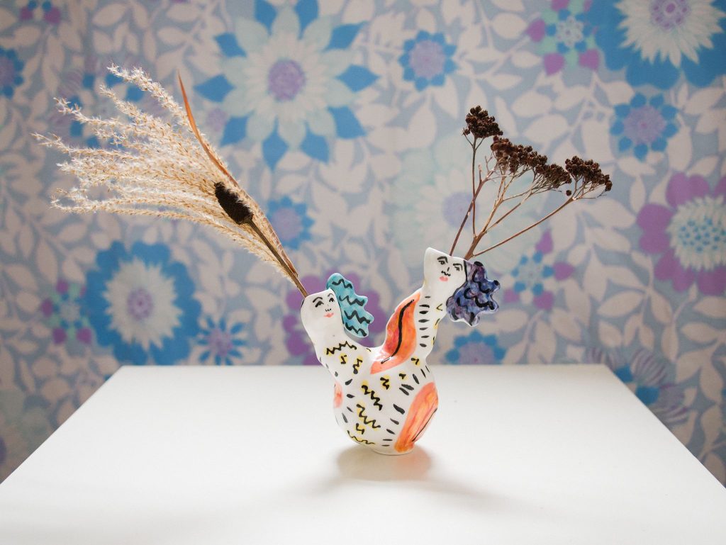 A vase sits on a white table with a blue patterned tablecloth background. The vase has to female heads splitting into different directions with dried flowers in each stem.